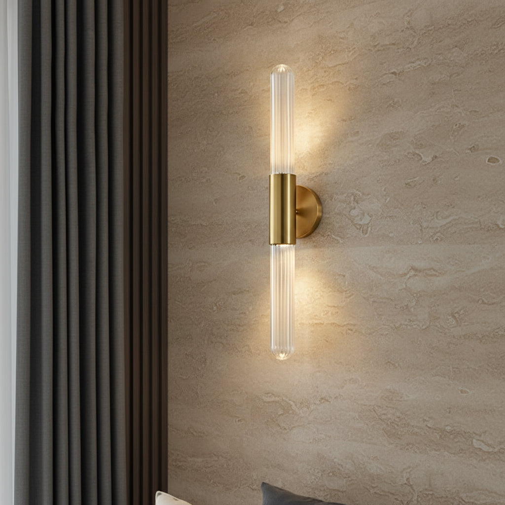 Minimalist wall light with clear fluted glass tube and brass backplate, lit warm.