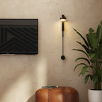 Black vertical wall sconce with cylindrical lamp head and pull-switch knob, glowing light